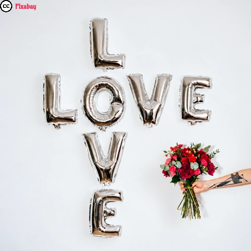 Silberne Luftballons bilden vor einer hellen Wand das Wort „LOVE“. Rechts ragt eine tätowierte Hand ins Bild und hält einen Strauß roter Rosen in Richtung des Schriftzugs.